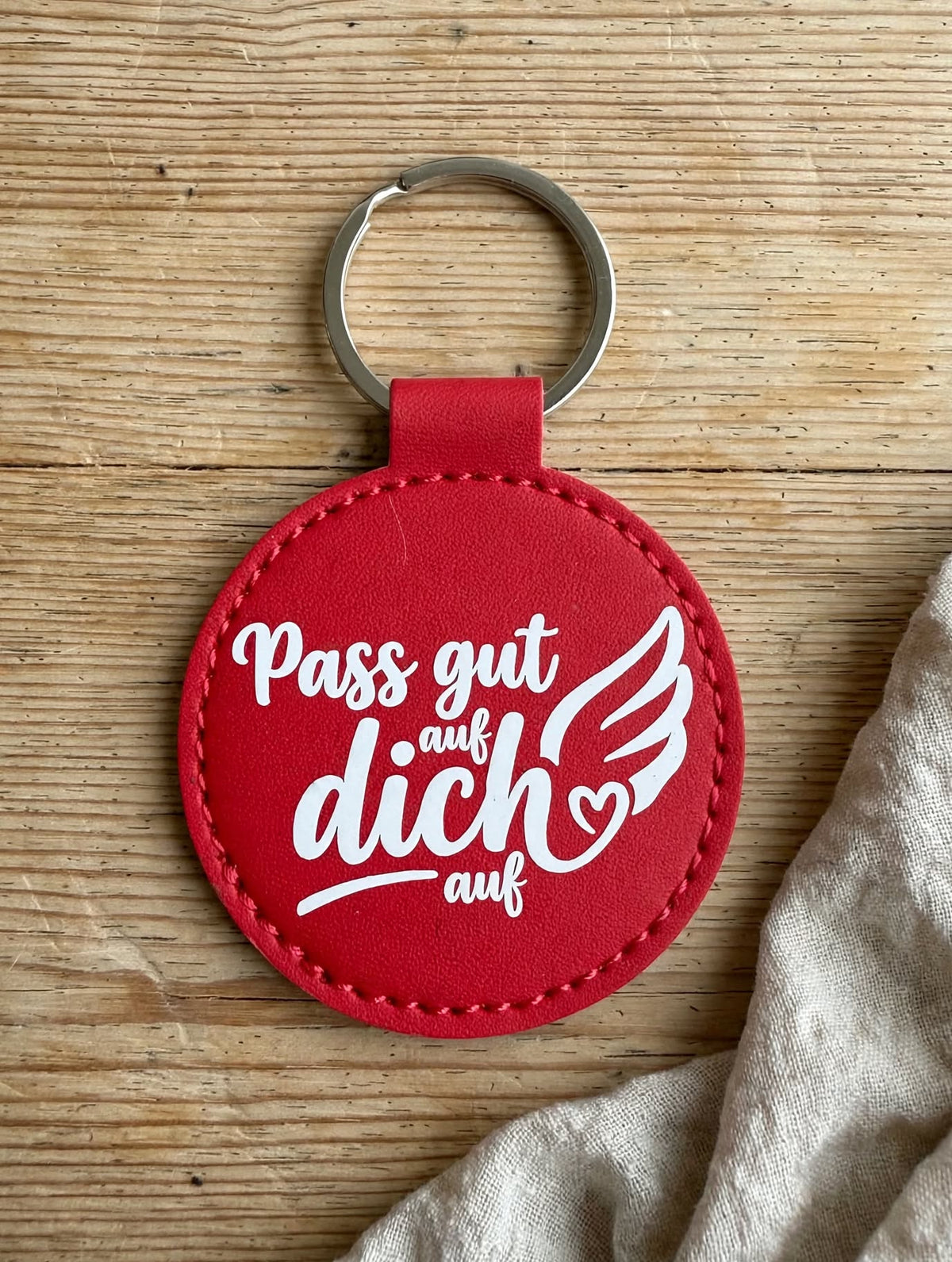 Schlüsselanhänger Pass gut auf dich auf rot - runder Kunstleder Anhänger mit weissem Spruch und Engelsflügel Herz - Detailansicht auf Holz - Geschenk Idee Schutzengel