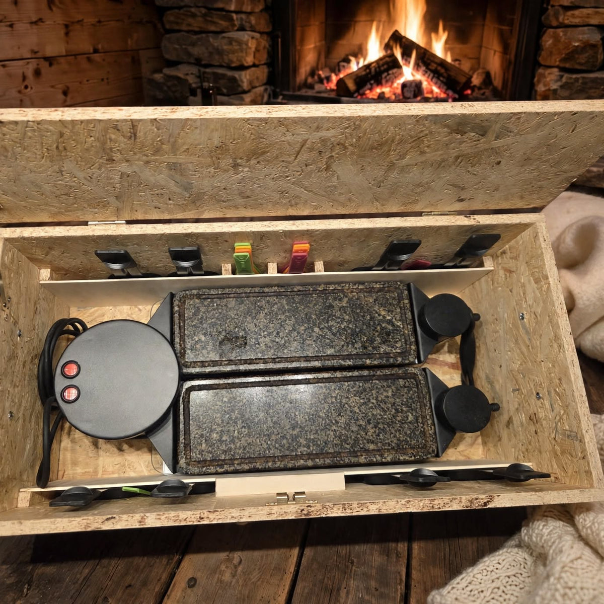 Raclette Ofen Kiste nach Mass offen mit heissem Stein und Pfännchen Fächern und Raclette Logo Berge Tannen vor Kaminfeuer mit Wolldecke, handgemacht OSB Holz, handgemacht in Hämikon bei Hitzkirch