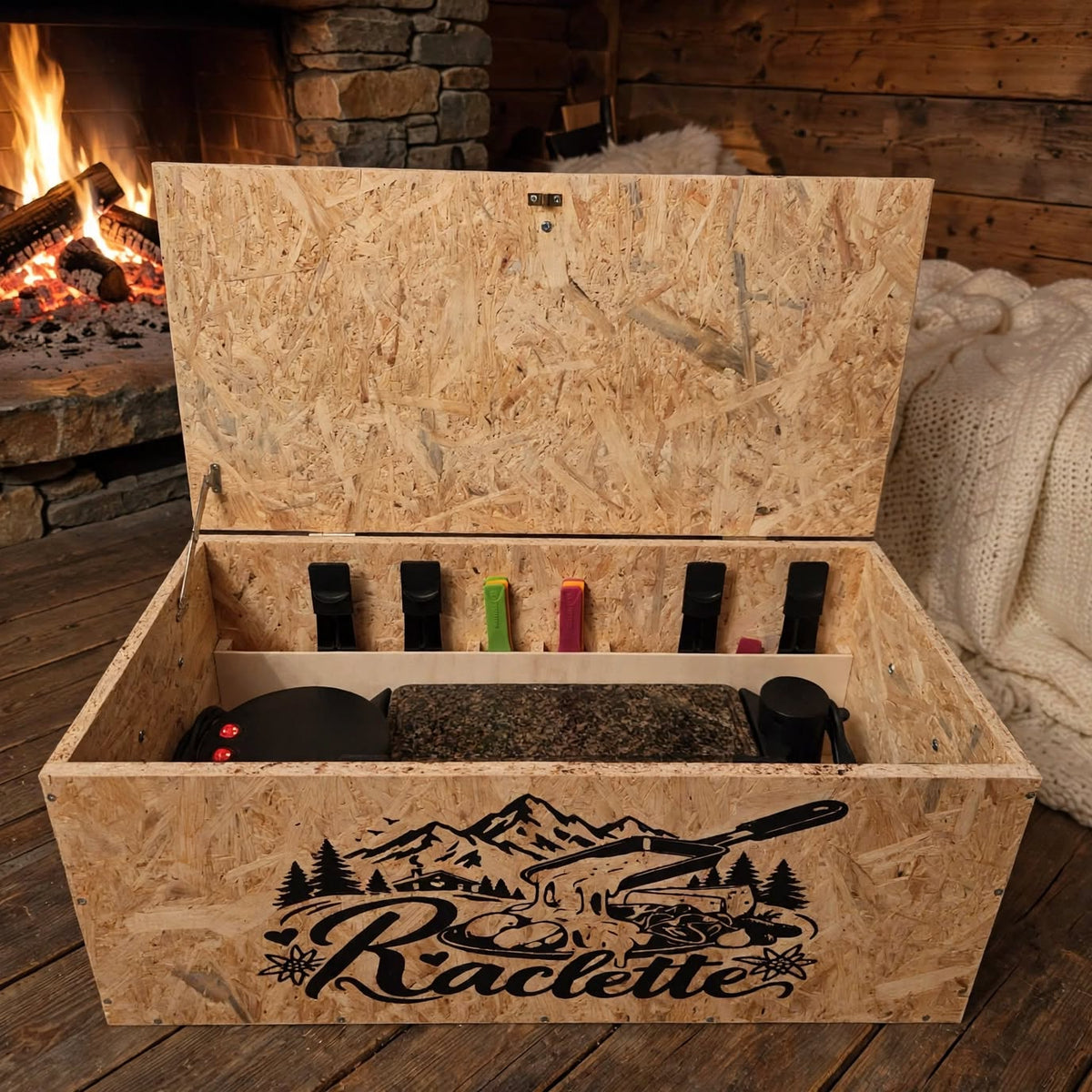 Raclette Ofen Kiste nach Mass offen mit heissem Stein und Pfännchen Fächern und Raclette Logo Berge Tannen vor Kaminfeuer mit Wolldecke, handgemacht OSB Holz, handgemacht in Hämikon bei Hitzkirch